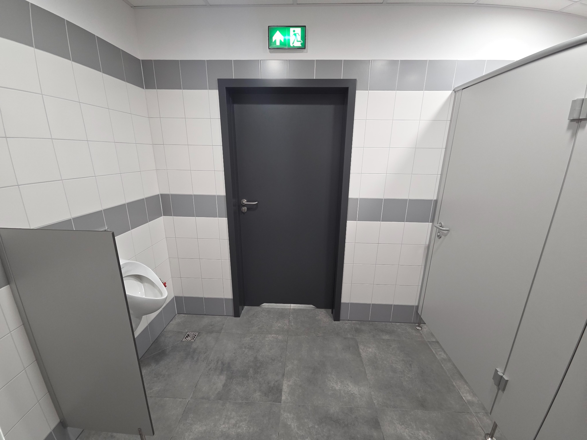 Wnętrze publicznej toalety z białymi i szarymi płytkami, szarą podłogą, pisuarami i kabinami. Nad drzwiami widoczny znak ewakuacyjny.