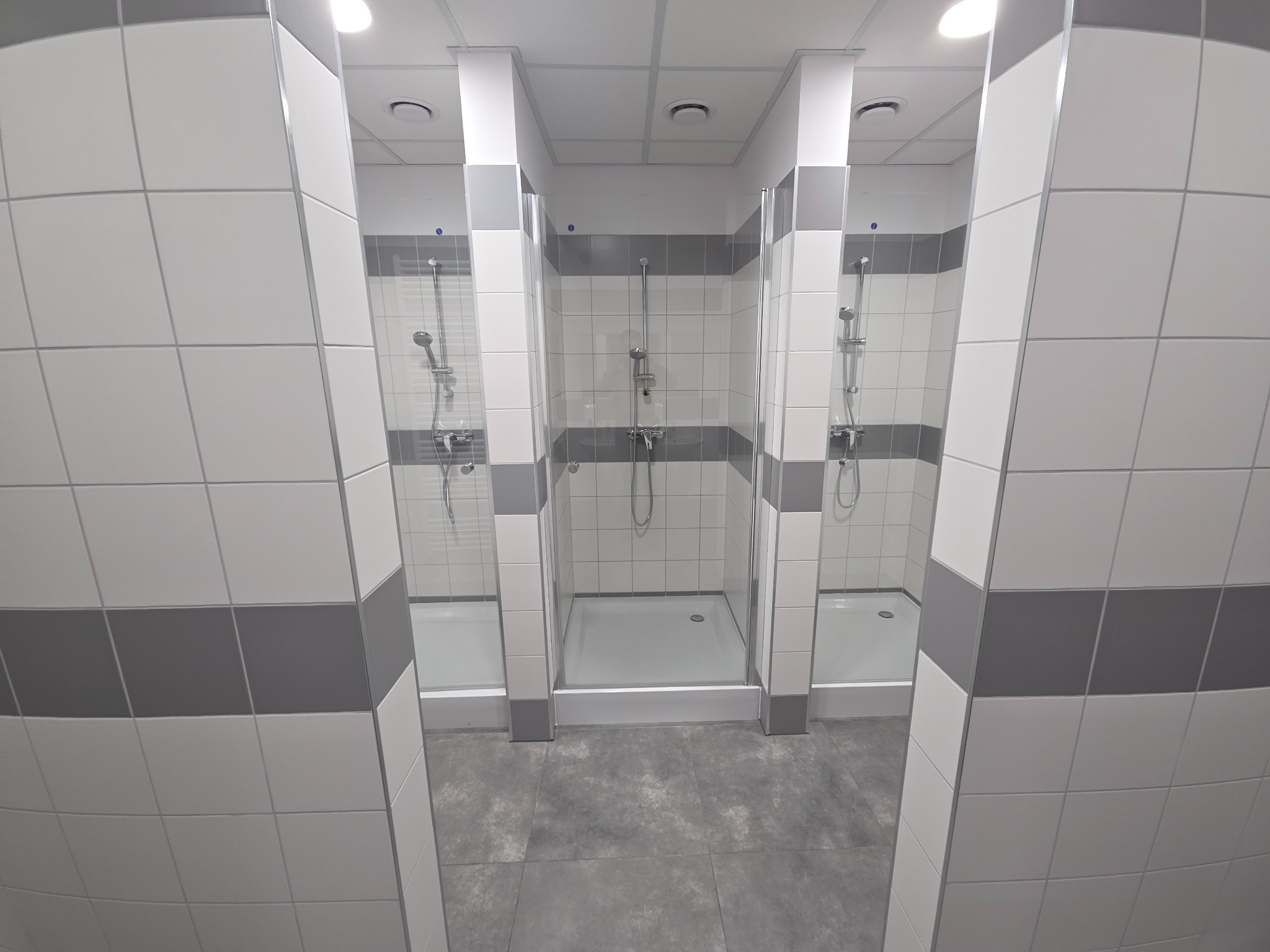 Trzy kabiny prysznicowe z białymi i szarymi płytkami, oddzielone ściankami. Widoczne słuchawki prysznicowe i brodziki. Szara podłoga z płytek.