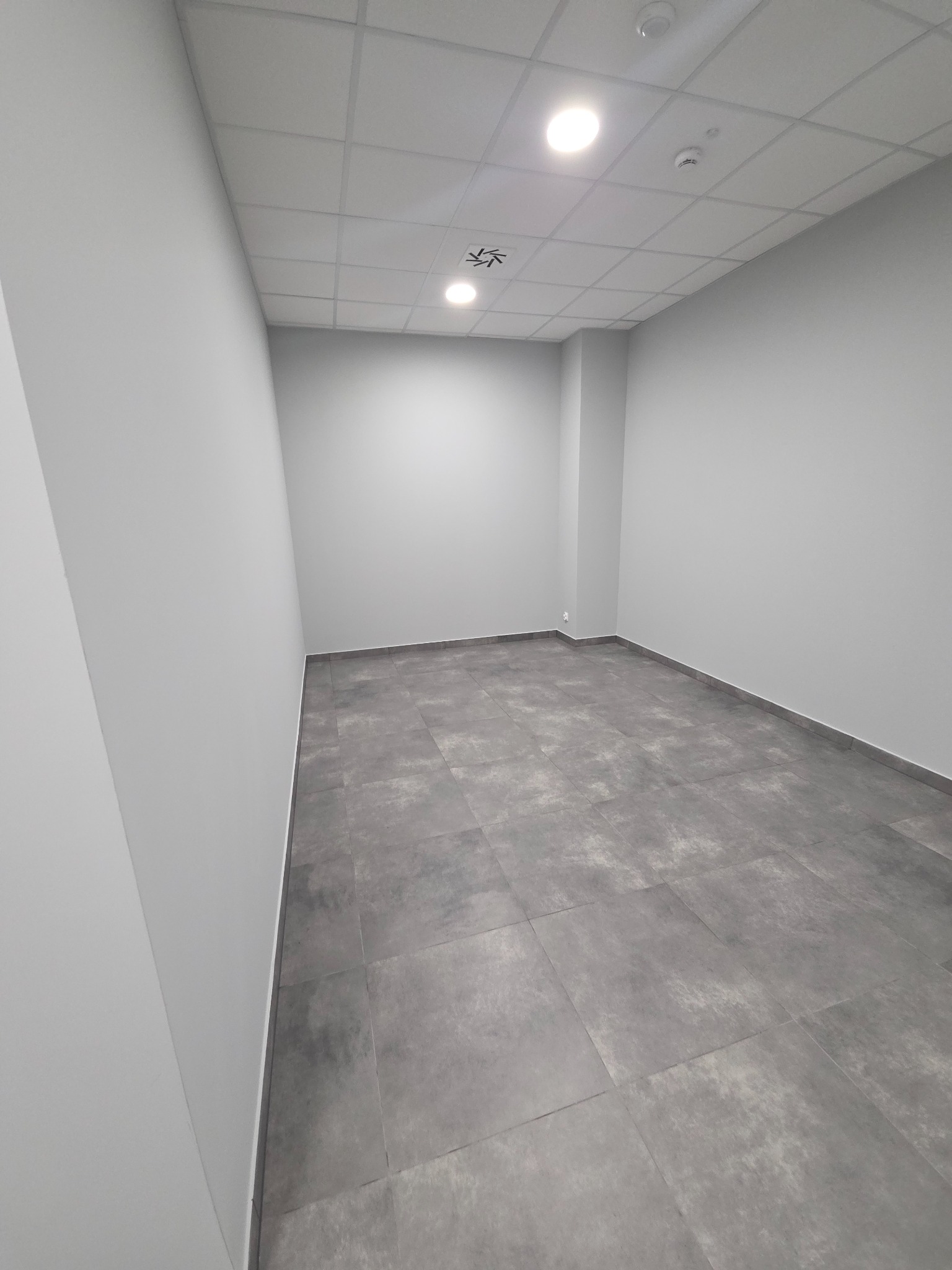 Wnętrze wykończonego, pustego pokoju z szarymi płytkami podłogowymi i białymi ścianami. Widoczne oświetlenie sufitowe i kratownica sufitowa.