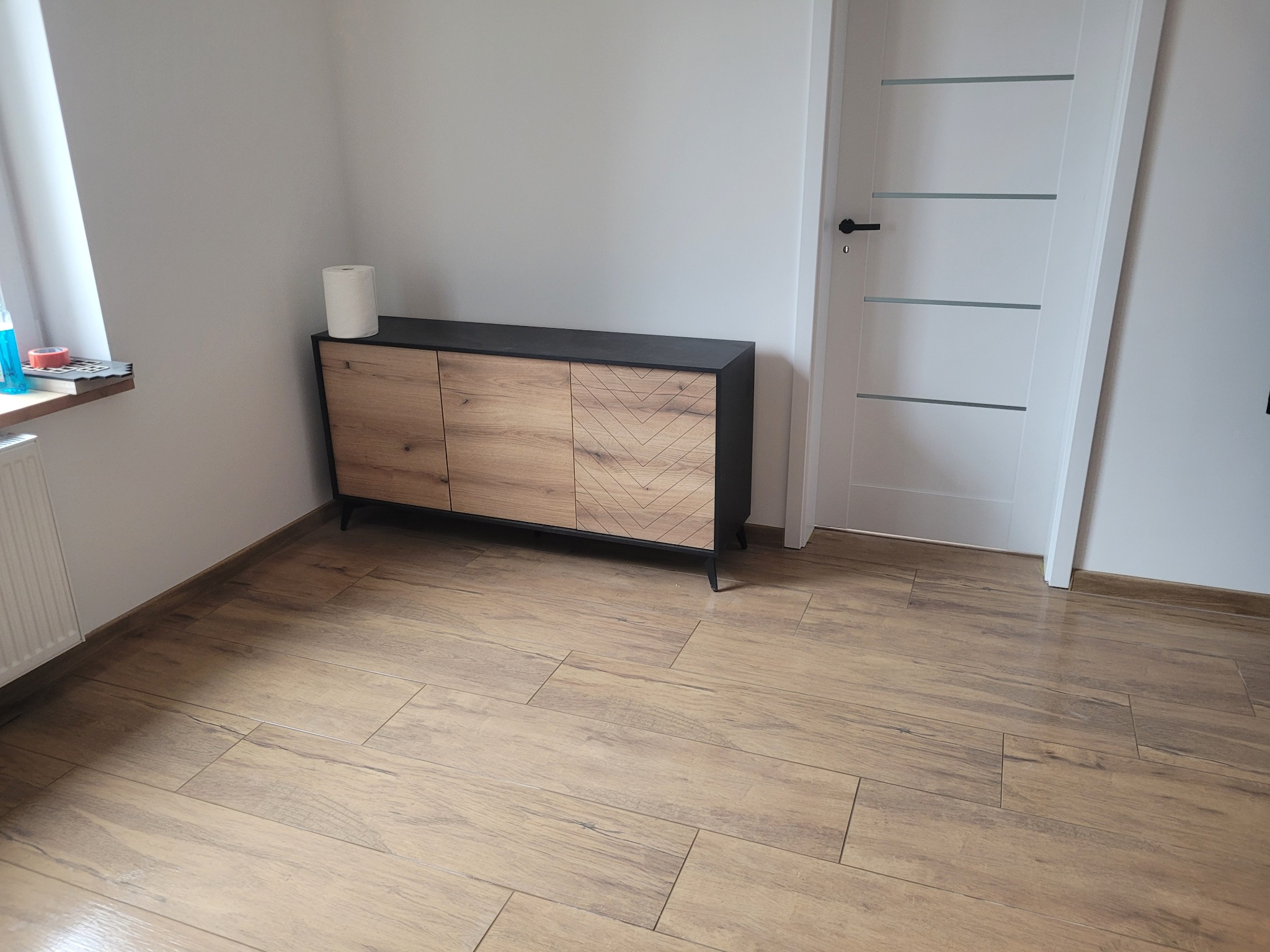 Salon z drewnopodobną podłogą z płytek, szafką RTV z dekorem jodełki i białą ścianą z drzwiami. Widok z góry, równomierne oświetlenie.