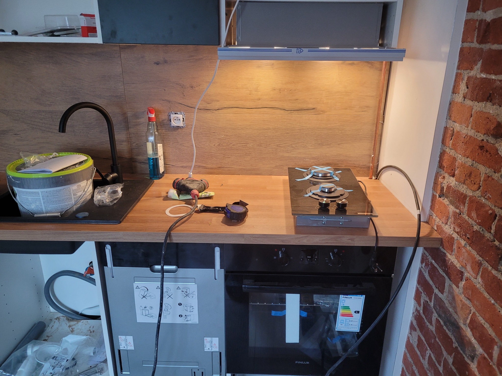 Montaż płyty gazowej i piekarnika w nowej kuchni, widoczne narzędzia i akcesoria montażowe, ściana z czerwonej cegły.