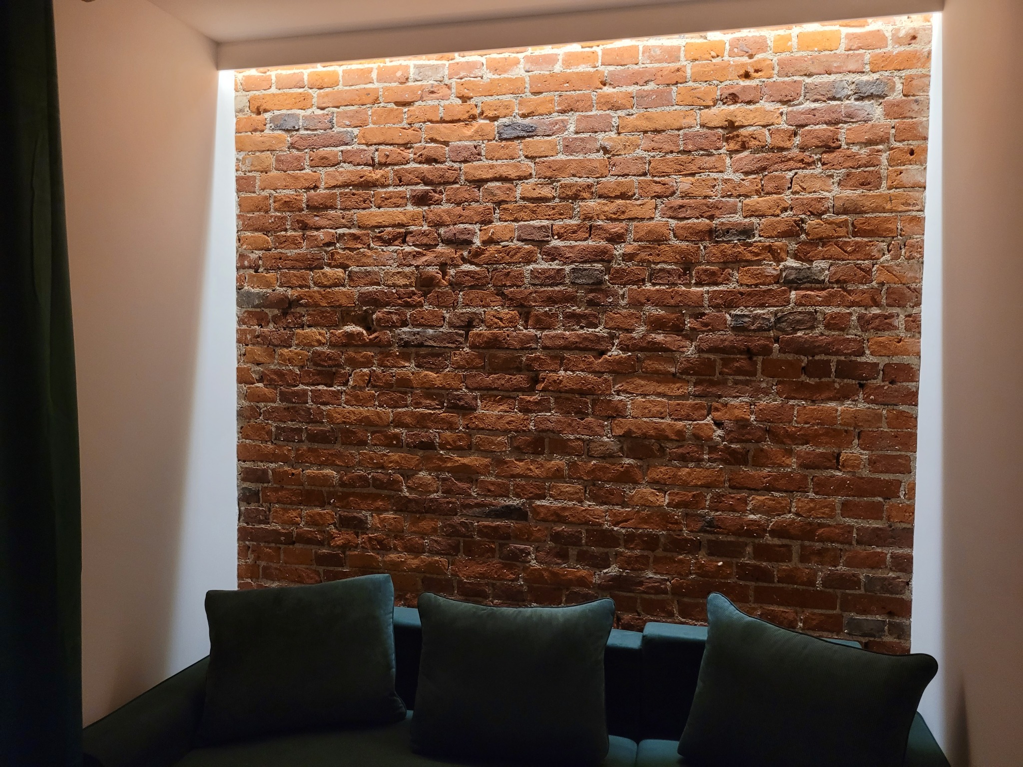 Ceglana ściana z podświetleniem LED w niszy, widok z dołu, fragment sofy z poduszkami w odcieniach zieleni. Dekoracyjne wykończenie wnętrza.