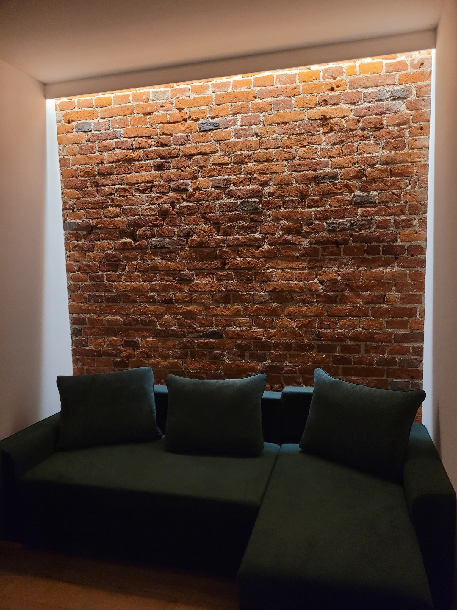 Ceglana ściana wnęki z oświetleniem, nad zieloną sofą z poduszkami. Widok na wykończone wnętrze z bliska. Minimalistyczny design z akcentem na teksturę cegły.