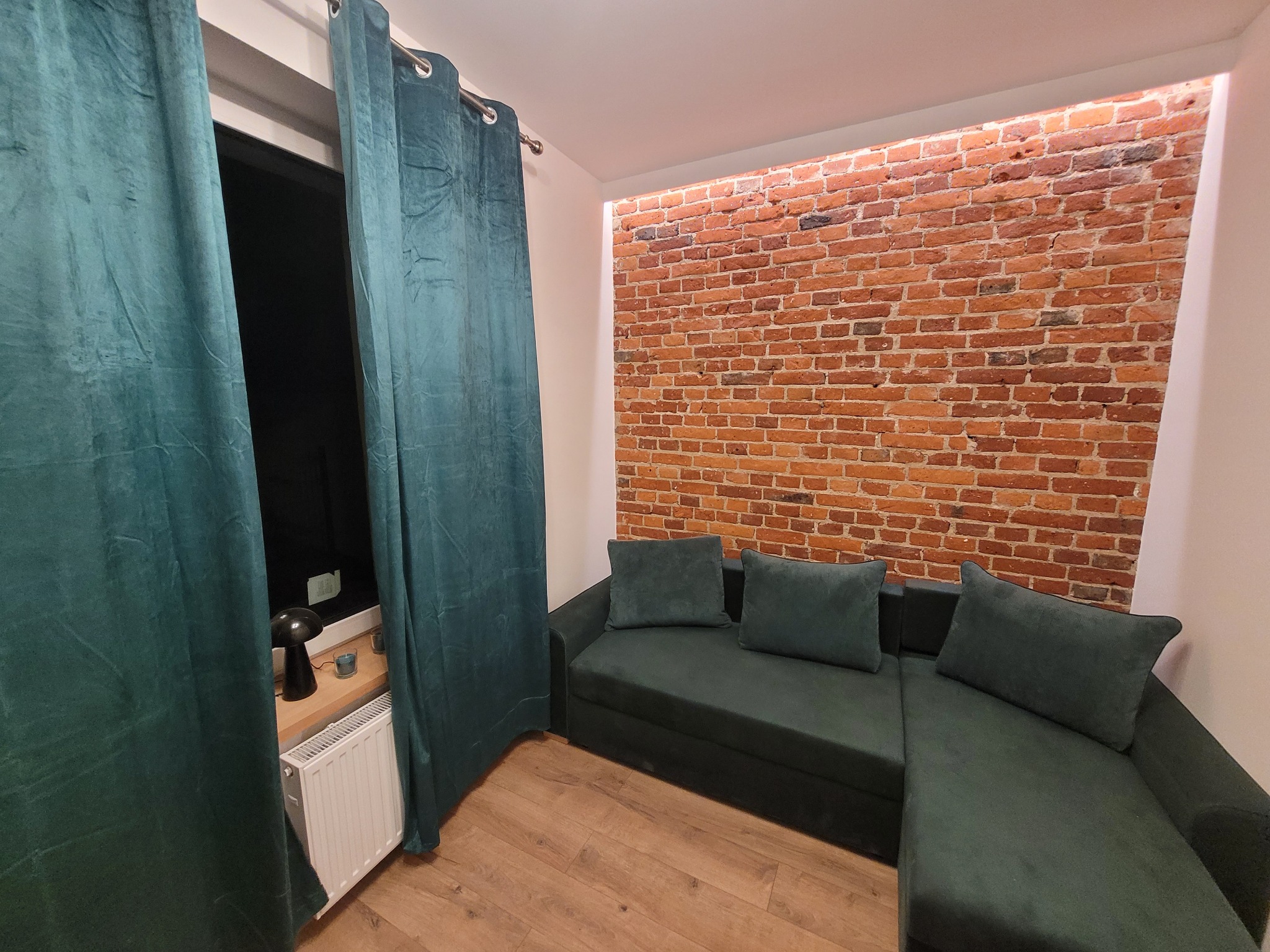 Salon z ceglaną ścianą i zieloną sofą. Widoczne okno z zasłonami i drewniana podłoga. Oświetlenie LED podkreśla strukturę cegieł.