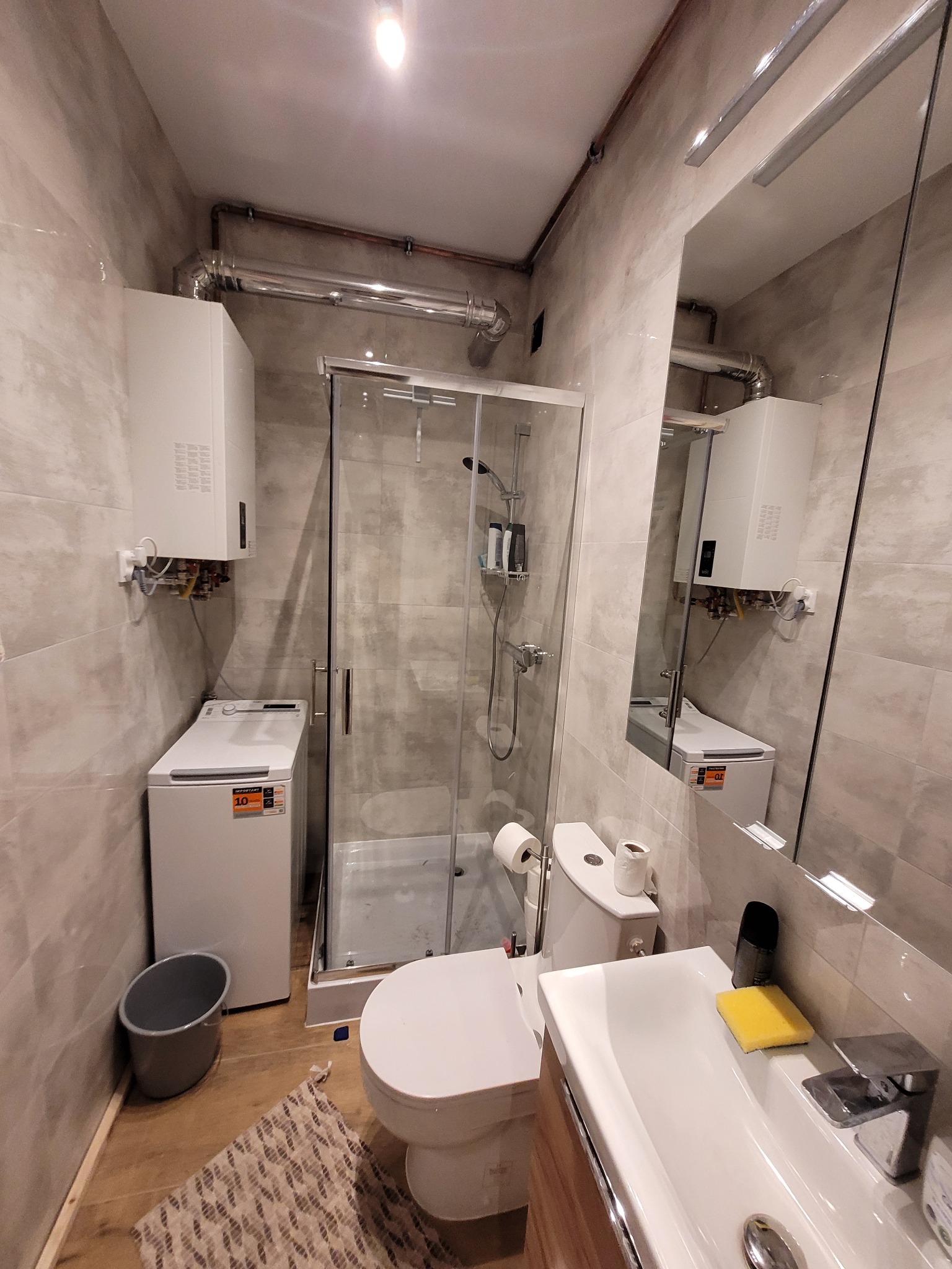 Jasna łazienka z kabiną prysznicową, umywalką i pralką. Widoczne rury pod sufitem. Ujęcie z niskiego kąta, podkreślające przestrzeń. Nowoczesny design.