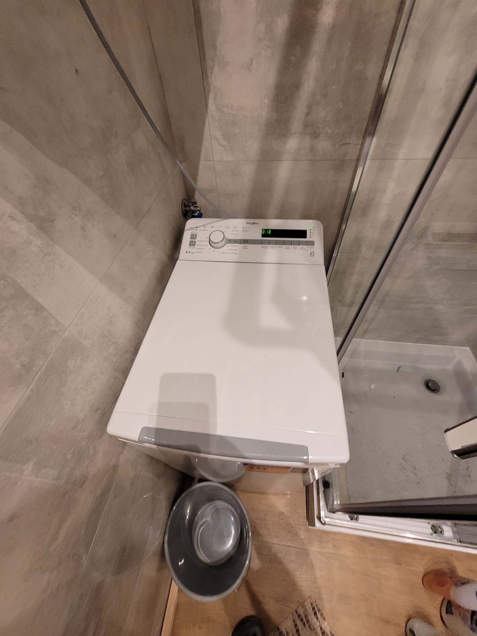 Biała pralka Whirlpool w rogu łazienki z szarymi płytkami, obok kabina prysznicowa. Ujęcie z góry, widoczny kosz na pranie.