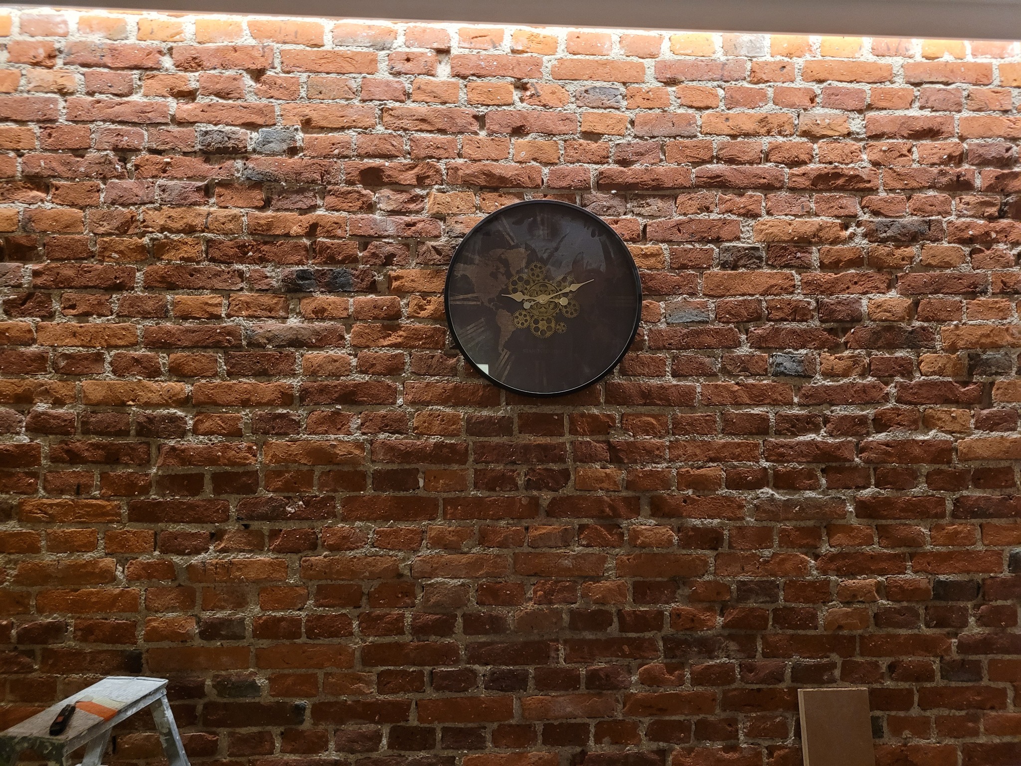 Ceglana ściana z okrągłym zegarem w stylu industrialnym. Widoczny fragment drabiny malarskiej. Oświetlenie LED podkreśla fakturę cegieł.