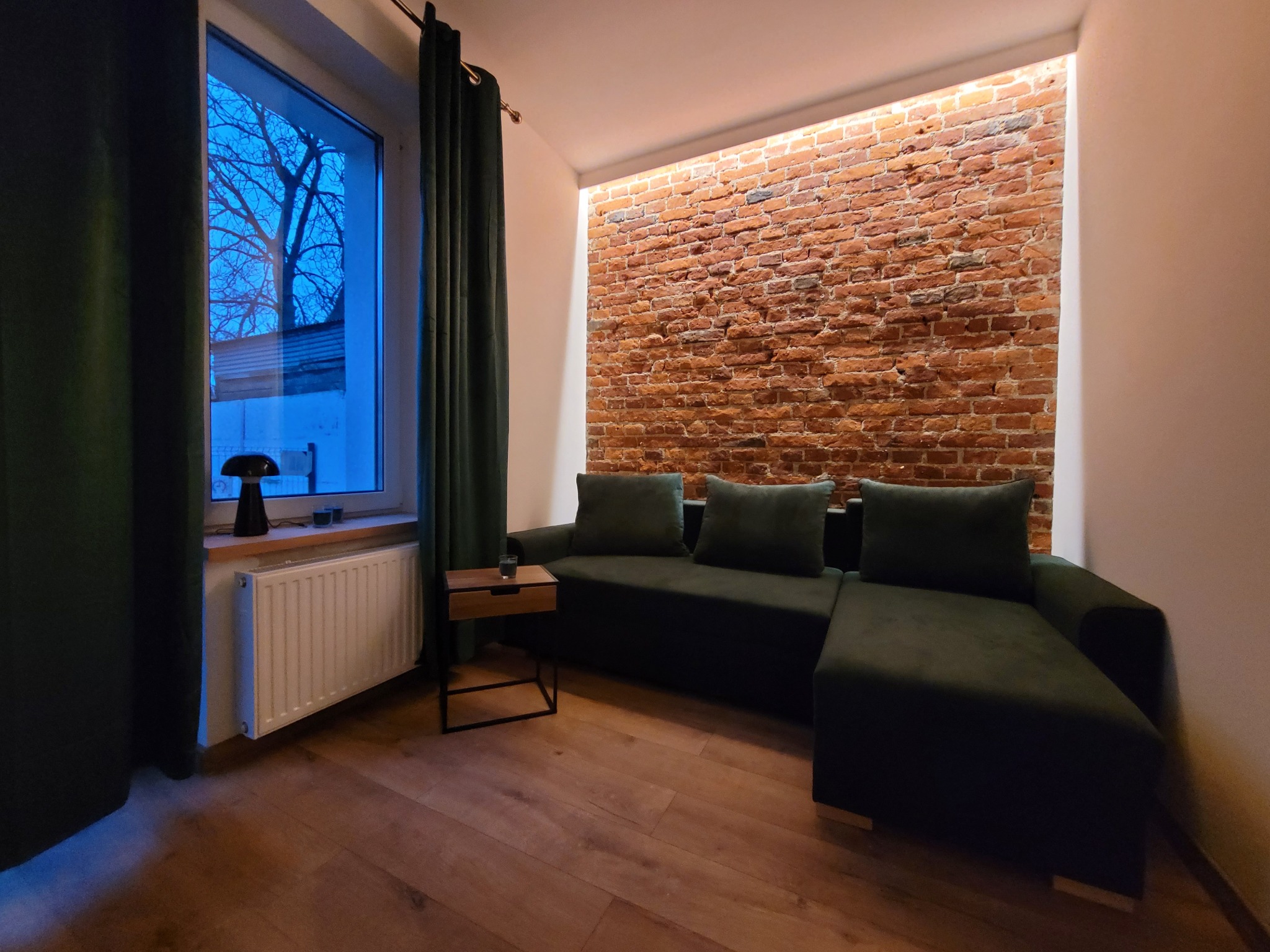 Salon z ceglaną ścianą, oświetleniem LED i zieloną sofą. Parkiet na podłodze, zasłony w oknie. Minimalistyczny stolik kawowy. Ciepłe, stonowane oświetlenie.