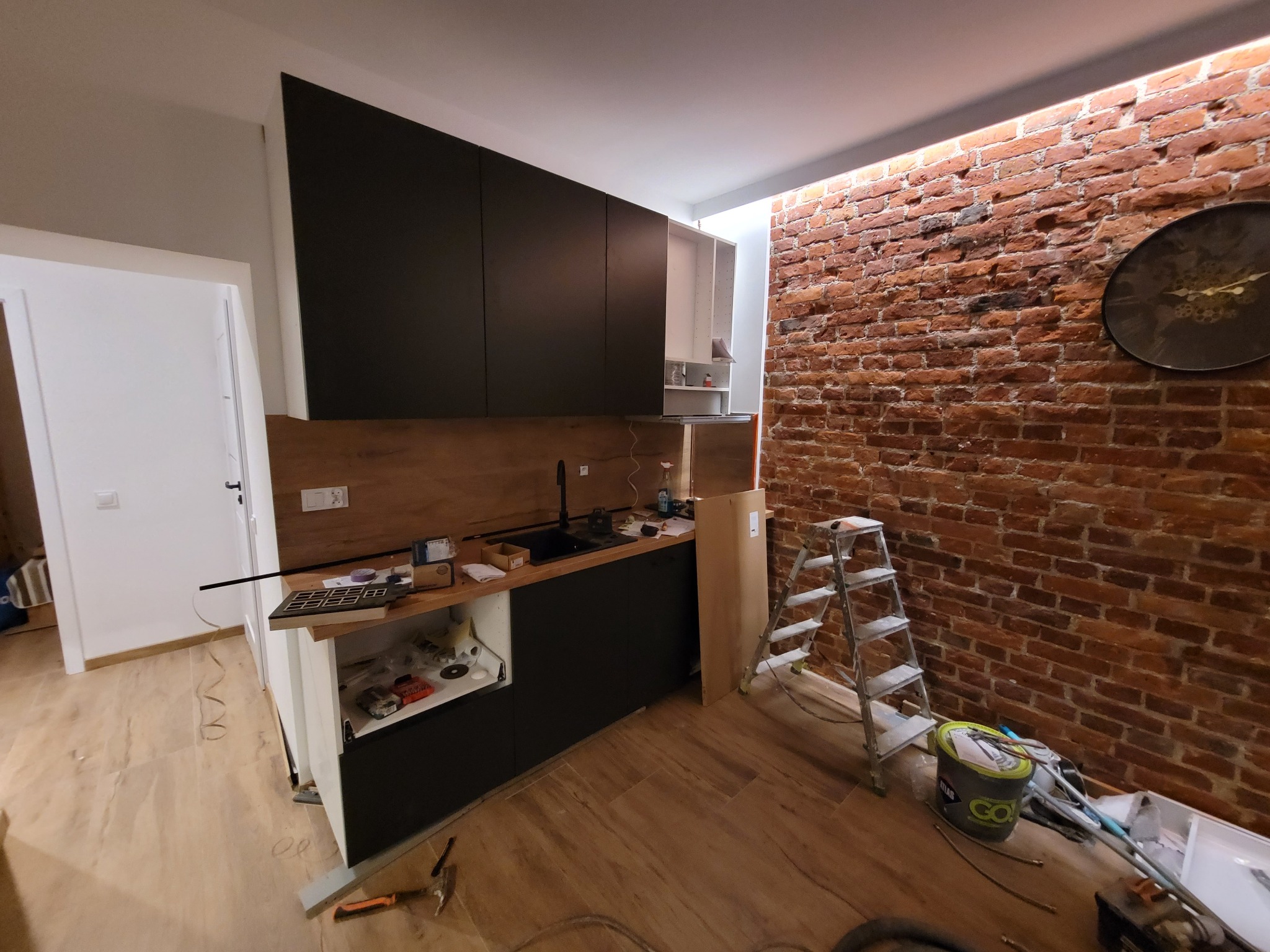 Montaż nowoczesnej, czarnej zabudowy kuchennej z drewnianym blatem na tle ściany z czerwonej cegły. Widoczna drabina i narzędzia, sugerujące etap wykończenia.