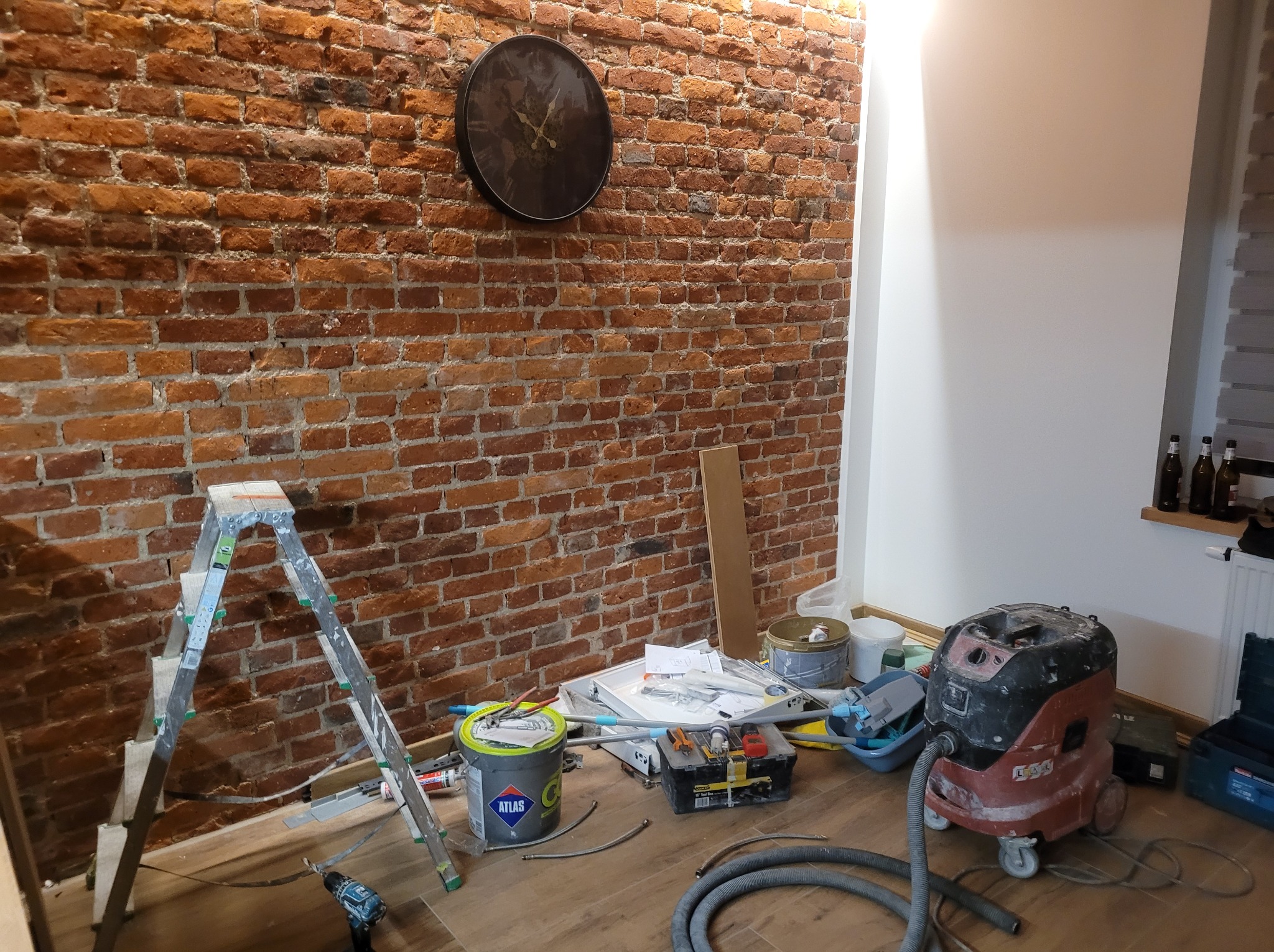 Ceglana ściana z zegarem w trakcie prac wykończeniowych. Widoczna drabina, narzędzia, odkurzacz przemysłowy i wiadro z klejem Atlas.