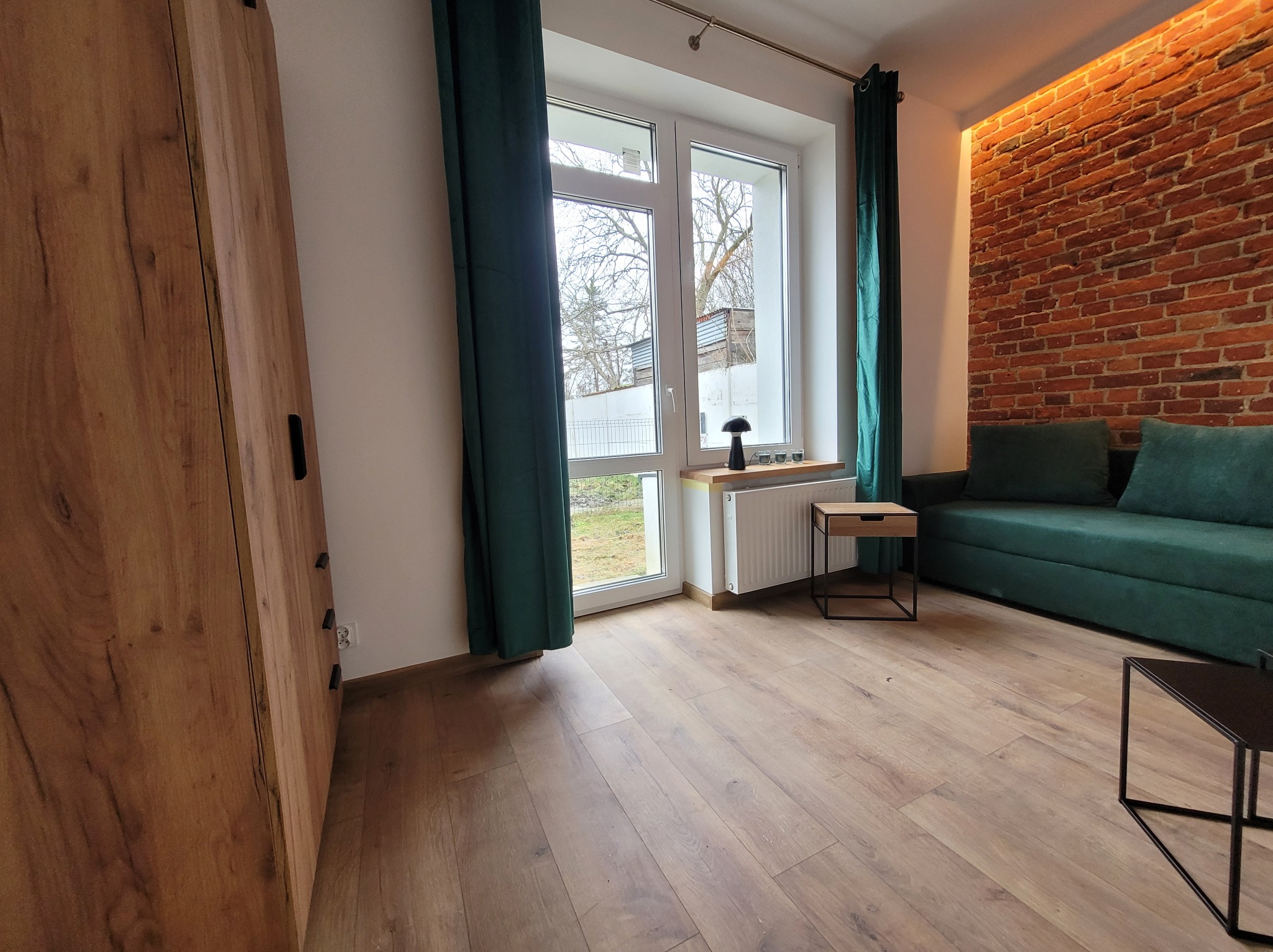 Jasne wnętrze pokoju z drewnianą podłogą, ceglaną ścianą z oświetleniem, zieloną sofą i oknem z zielonymi zasłonami. Widoczny fragment szafy.
