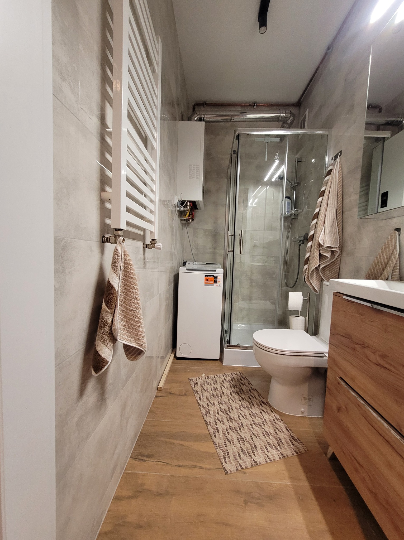 Wykończona łazienka w Węgorzynie z kabiną prysznicową, toaletą, umywalką z szafką z drewnianym frontem i grzejnikiem drabinkowym. Na podłodze leży dywanik.