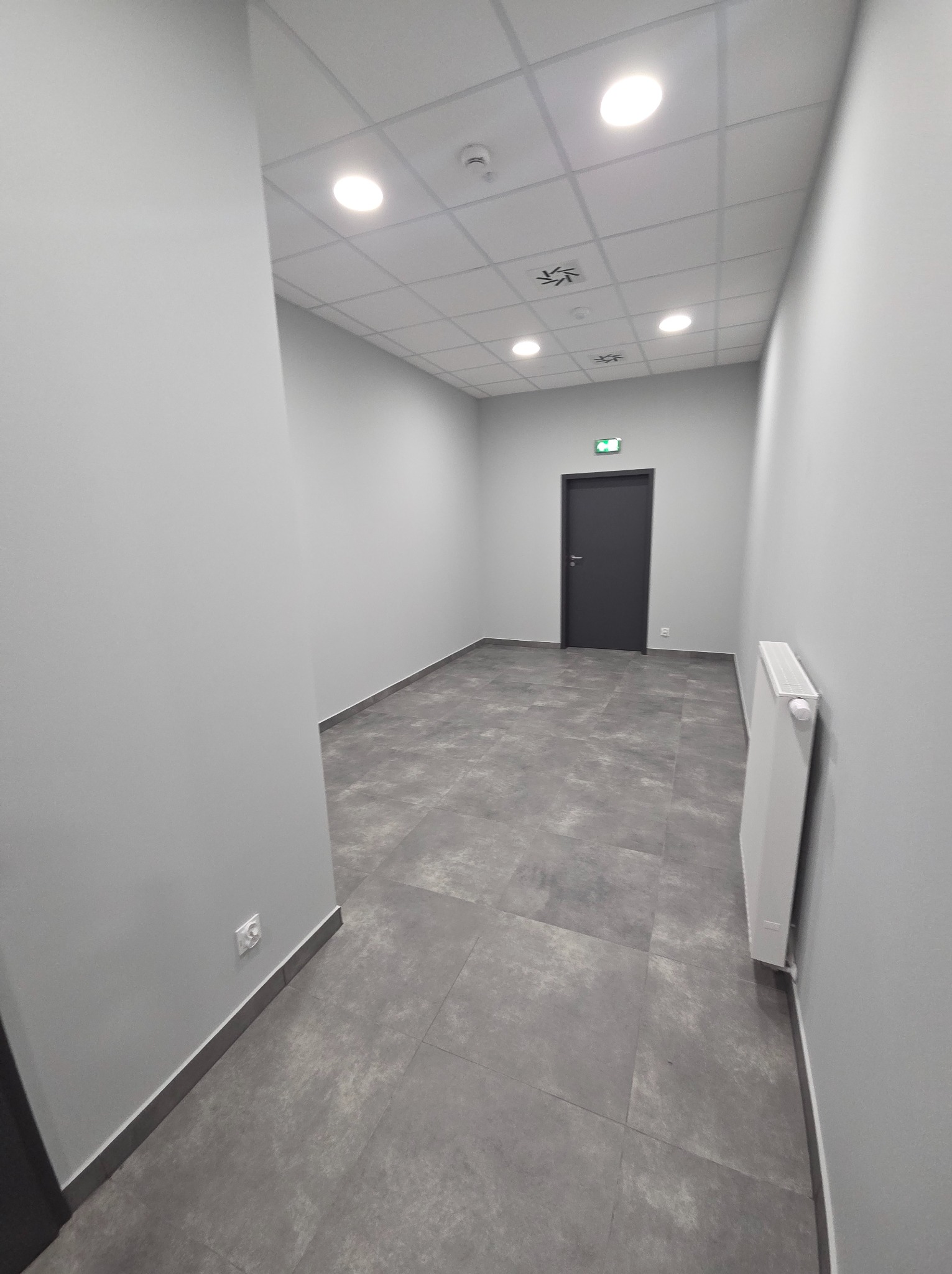 Szare płytki podłogowe w korytarzu biurowym, widoczny grzejnik i drzwi ewakuacyjne. Minimalistyczne wykończenie wnętrza z oświetleniem sufitowym.
