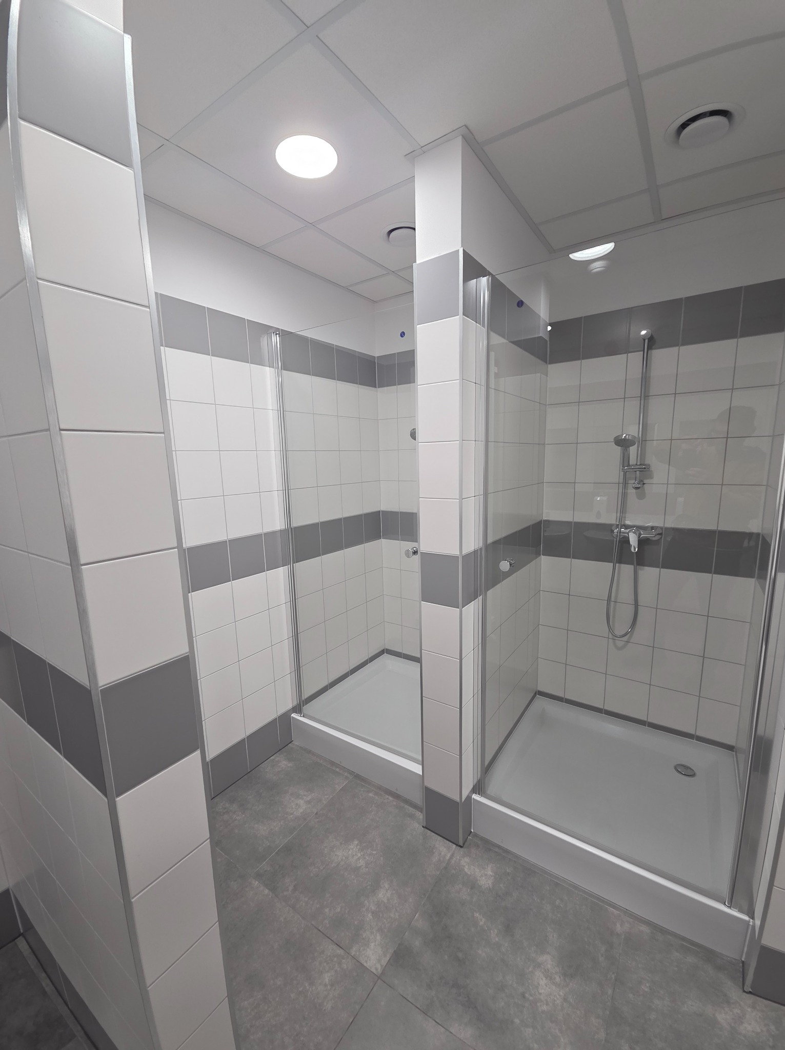 Nowoczesna łazienka publiczna z kabinami prysznicowymi wykończonymi białymi i szarymi płytkami, szklane drzwi, szara podłoga, oświetlenie sufitowe.
