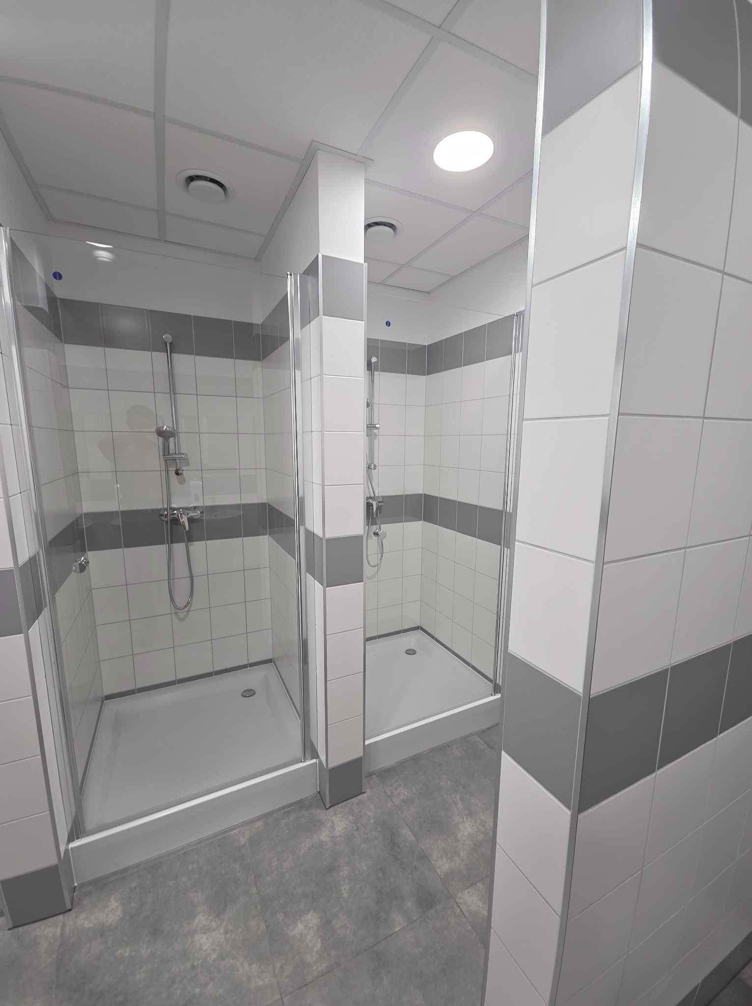 Dwie kabiny prysznicowe z szarymi akcentami w płytkach, oddzielone ścianką. Widoczne brodziki i armatura. Oświetlenie sufitowe.