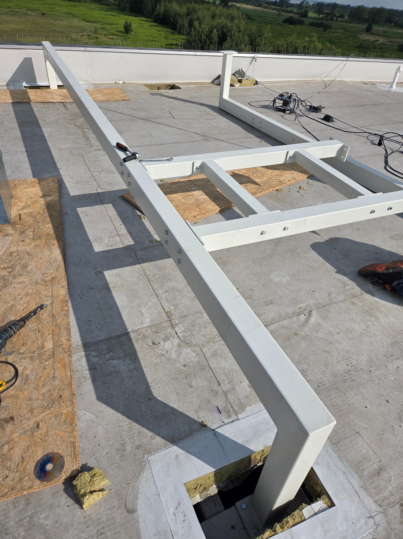 Biała, metalowa konstrukcja na płaskim dachu z widocznymi narzędziami i elementami montażowymi. W tle zielony krajobraz.