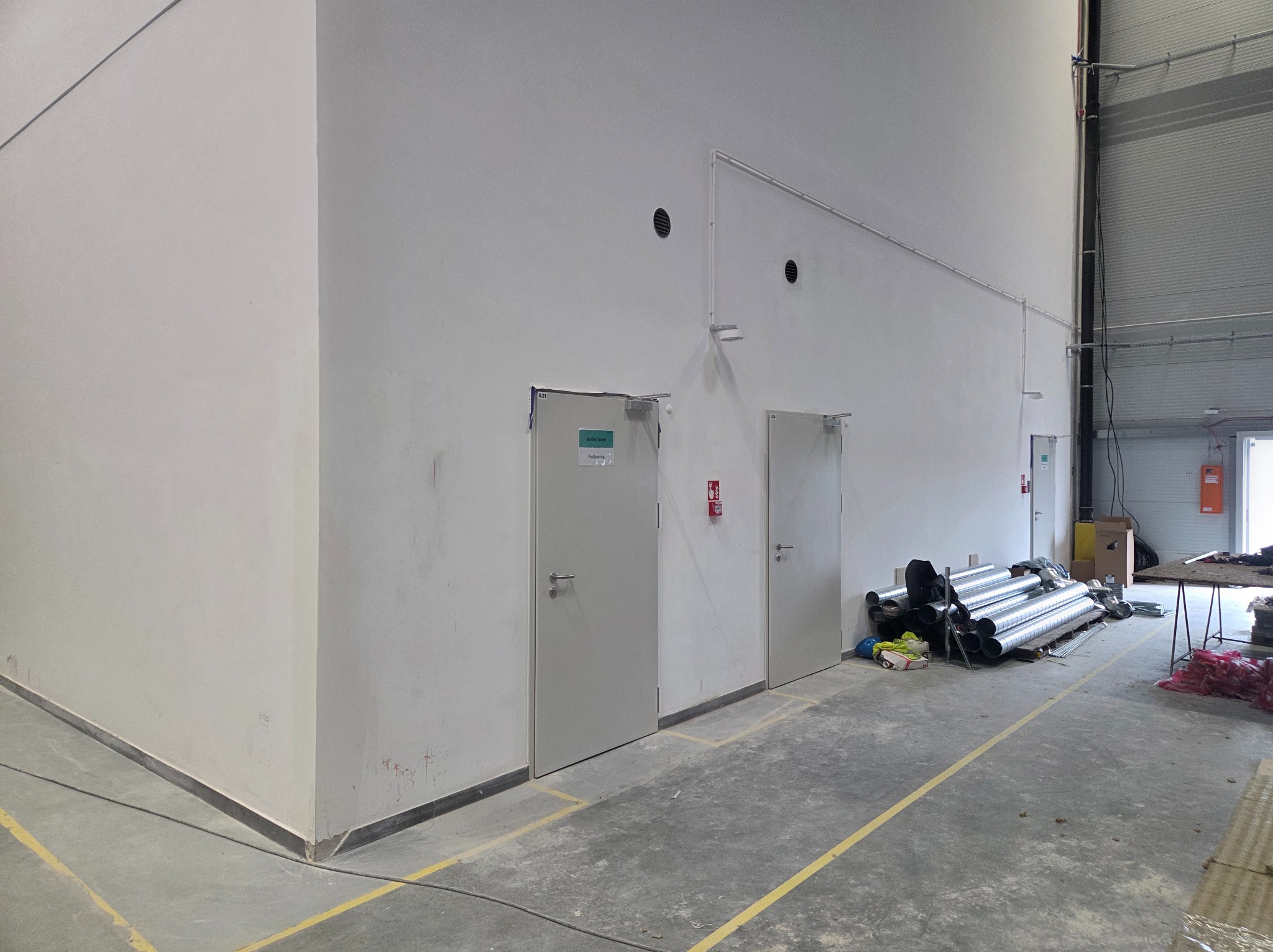 Wnętrze hali w trakcie wykończenia: białe ściany z trzema drzwiami, widoczne rury wentylacyjne i materiały budowlane na szarej posadzce z żółtymi liniami.