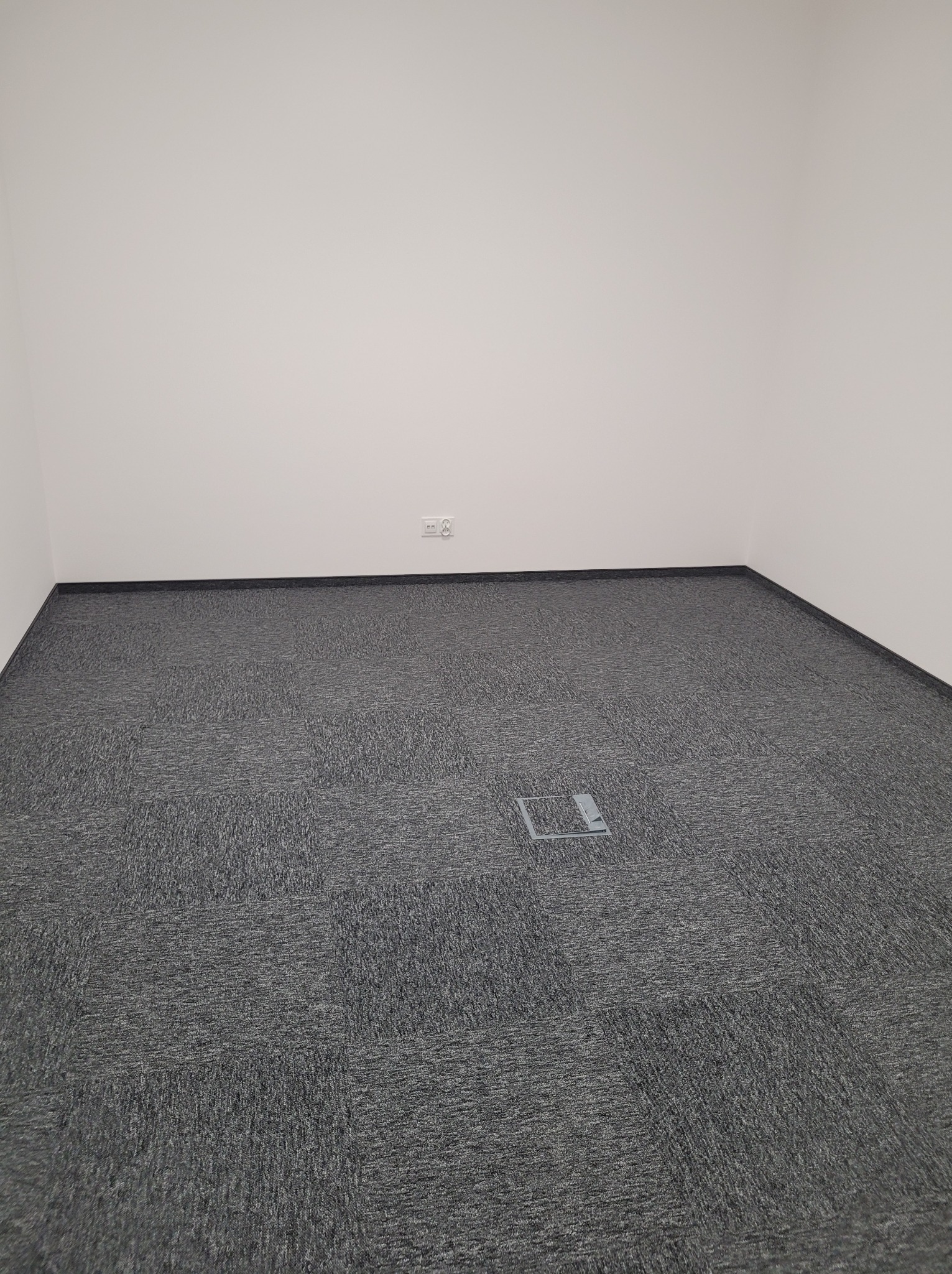 Wnętrze biurowe po wykończeniu: szara wykładzina dywanowa w kwadraty, białe ściany, gniazdko elektryczne i metalowa puszka podłogowa. Minimalistyczny design.