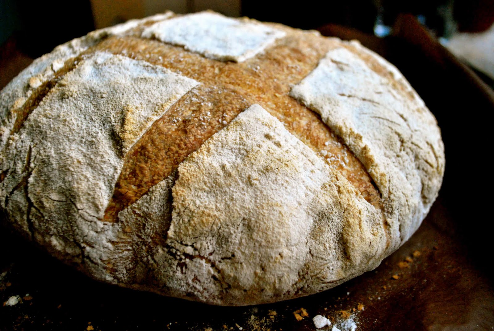 Okrągły bochenek chleba rustykalnego z popękaną skórką, posypany mąką w kształcie krzyża, leżący na ciemnym drewnianym podłożu.