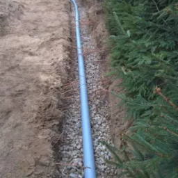 Szary rurociąg ułożony w wykopie wypełnionym drobnymi kamieniami, obok rzędu zielonych drzew iglastych, na działce z piaszczystą ziemią.