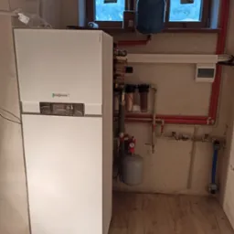 Biała pompa ciepła Viessmann Vitocal 222 z niebieskim zbiornikiem wyrównawczym, instalacja hydrauliczna z filtrami i manometrem, zamontowana w pomieszczeniu z dwoma oknami