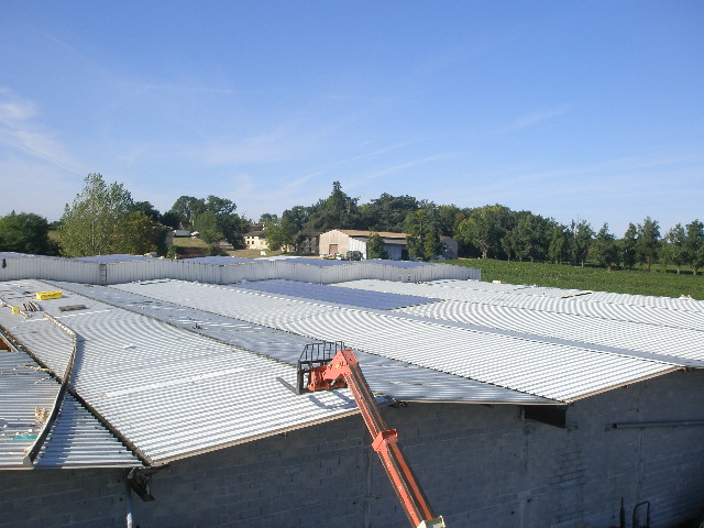 Widok z góry na rozległy dach hali z blachy falistej w trakcie montażu, z podnośnikiem koszowym w prawym dolnym rogu i wiejską scenerią w tle.