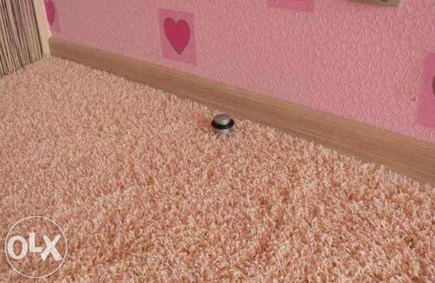 Różowy dywan shaggy z widocznym ogranicznikiem do drzwi na tle ściany w serduszka i drewnianej listwy przypodłogowej. Ciepłe oświetlenie.