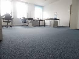 Wnętrze szwalni z niebieską wykładziną dywanową. Widoczne biurka z maszynami do szycia i krzesłami. Jasne, przestronne pomieszczenie o minimalistycznym wystroju.