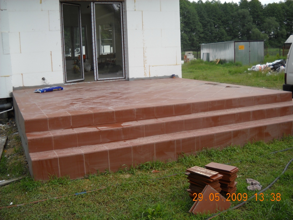 Wykończony taras z brązowych płytek z trzema stopniami przed budynkiem w trakcie budowy, widoczne paczki płytek na trawie.