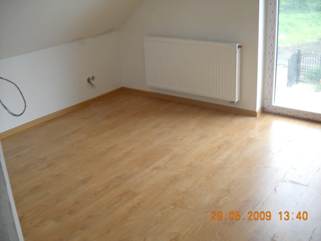 Pokój na poddaszu z nową, jasną podłogą imitującą drewno, grzejnikiem na ścianie i oknem balkonowym z widokiem na zieleń. Widać wystające kable elektryczne ze ściany.