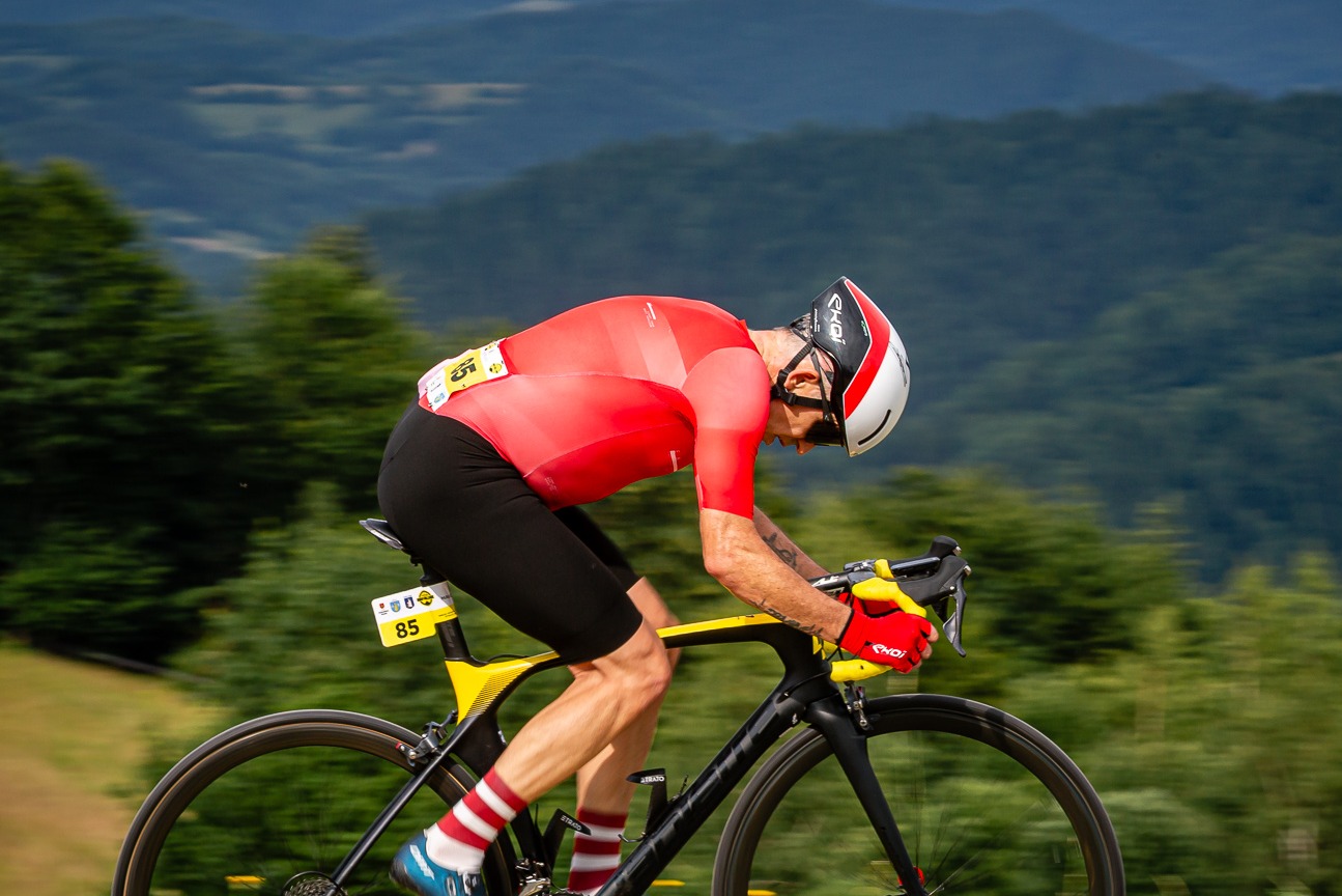 Kolarz w czerwonej koszulce i kasku, pochylony nad czarno-żółtym rowerem, jedzie pod górę na tle zielonych wzgórz. Numer startowy 85 na ramie roweru.