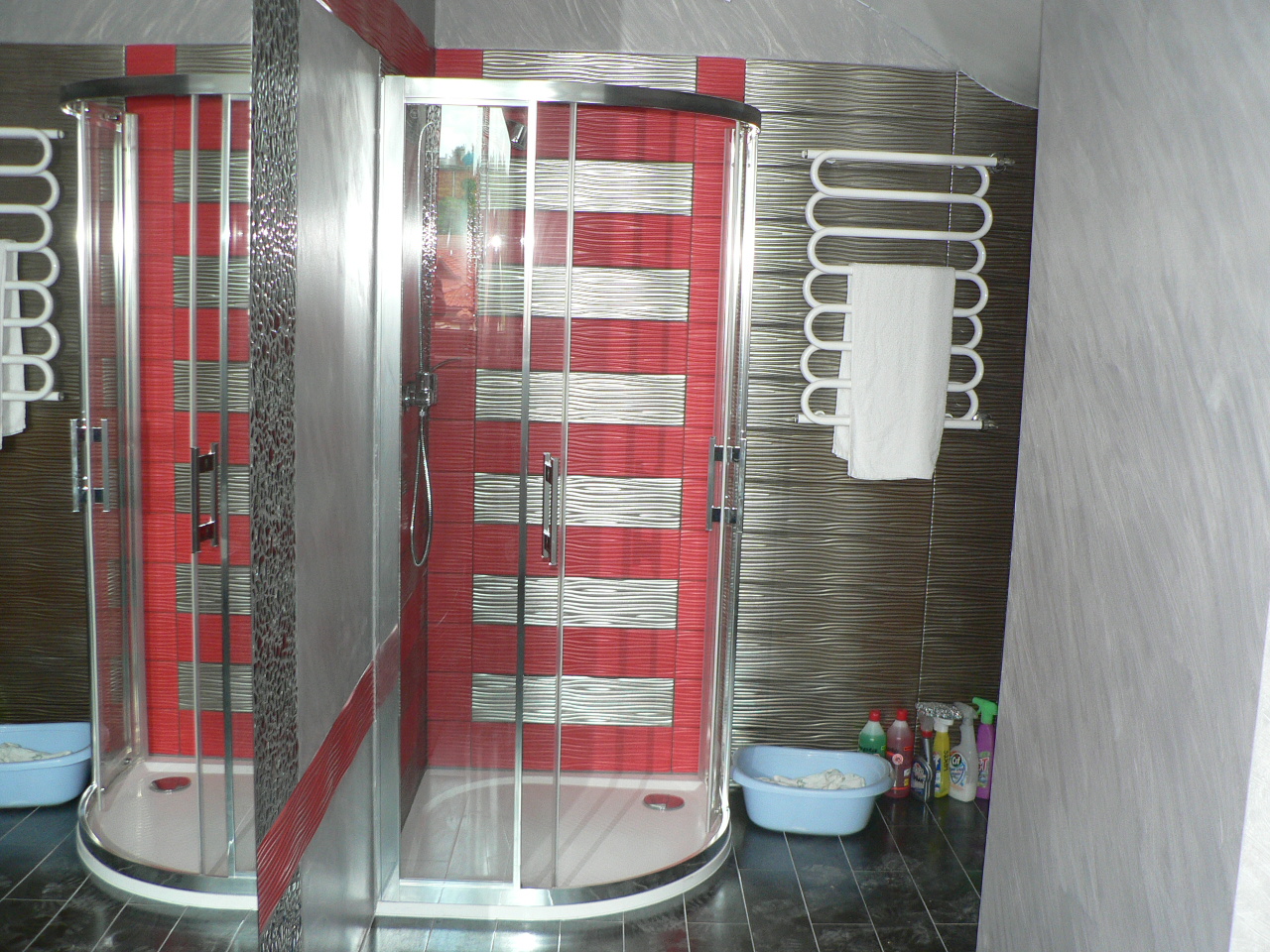 Dwie kabiny prysznicowe z czerwono-szarymi płytkami w paski, biały grzejnik drabinkowy z ręcznikiem, oraz środki czystości w niebieskiej misce na podłodze z ciemnych płytek.