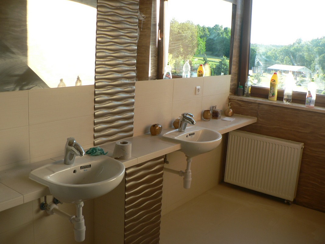 Jasna łazienka z dwoma umywalkami, dekoracyjnymi panelami ściennymi o falistej strukturze i widokiem na zieleń za oknem.