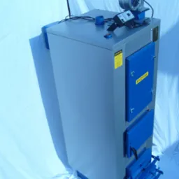 Nowy, szaro-niebieski kocioł na paliwo stałe z wentylatorem i panelem sterowania na górze, sfotografowany na białym tle.