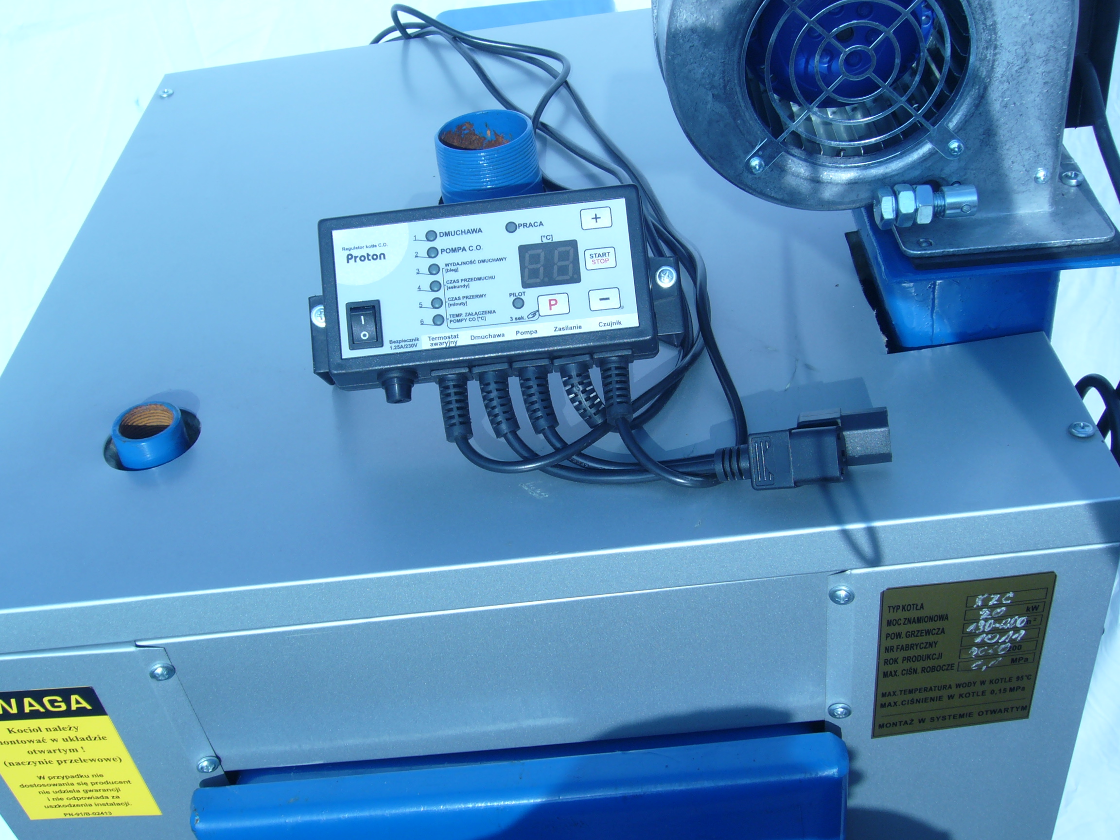 Szary kocioł z zamontowanym regulatorem Proton, widoczny wentylator, przewody i tabliczka znamionowa z parametrami technicznymi.