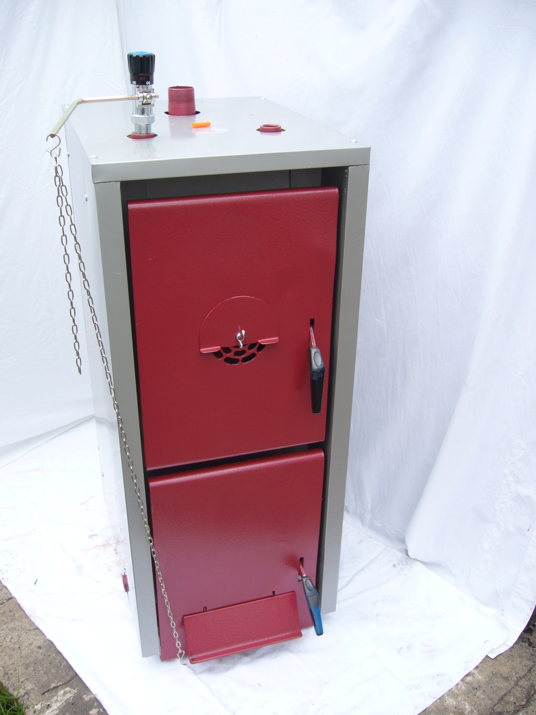 Nowy, dwudrzwiowy kocioł na paliwo stałe, z łańcuchem i zaworem regulacyjnym na górze, ustawiony na białym płótnie.