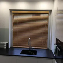 Kuchnia z drewnianymi żaluzjami poziomymi w oknie, czarny blat i białe szafki.