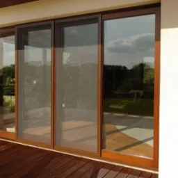 Przesuwne drzwi balkonowe z moskitierą w brązowej ramie, widok z zewnątrz na drewniany taras i ogród.