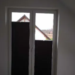 Ciemne plisy zamontowane w oknie dwuskrzydłowym, widok na dach sąsiedniego domu i zalesione wzgórze w tle.