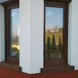 Dwa okna z brązowymi ramami i zwiniętymi, brązowymi osłonami zewnętrznymi w białej elewacji z ceglanym parapetem, widok z zewnątrz.