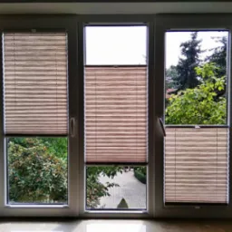 Trzyczęściowe okno z zamontowanymi plisowanymi osłonami okiennymi w kolorze beżowym, częściowo zasłonięte, widok na zieleń ogrodu i ścieżkę.