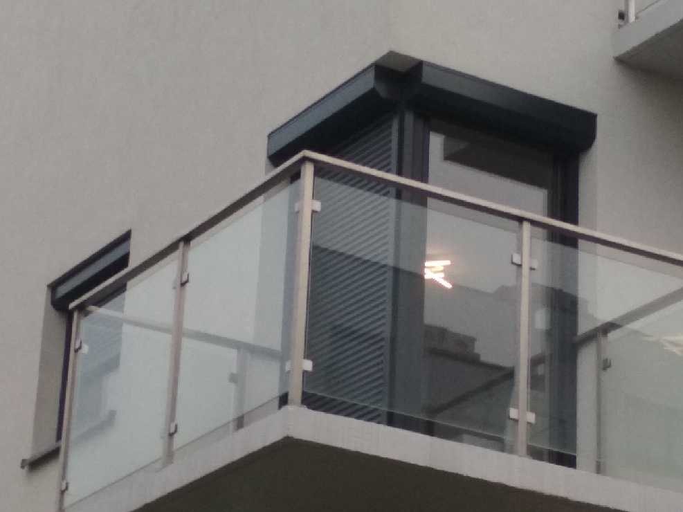 Balkon z szklaną balustradą i częściowo opuszczoną, grafitową przesłoną okienną, widoczną przez szybę, odbijającą światło.