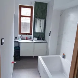 Nowoczesna łazienka po remoncie z białą wanną, umywalką i zielonymi płytkami na ścianie, widok zza futryny drzwi.