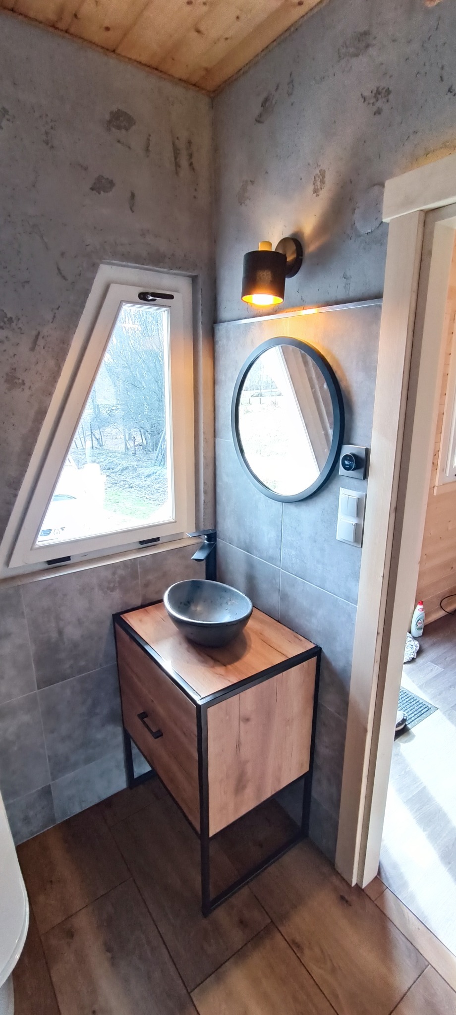 Nowoczesna łazienka z umywalką nablatową, szafką z drewnianymi frontami i czarną ramą, okrągłym lustrem z czarną obwódką, lampą ścienną, i oknem o nietypowym trójkątnym kształcie.