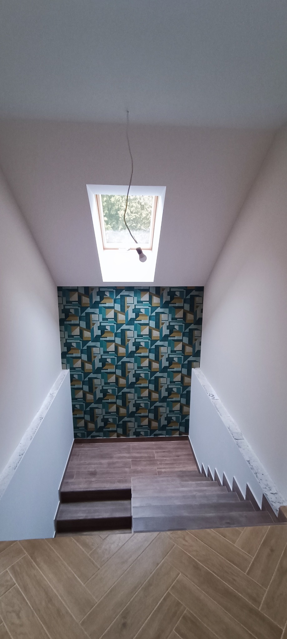 Klatka schodowa w trakcie wykańczania, z geometryczną tapetą w odcieniach turkusu i brązu na ścianie, widoczne schody wyłożone płytkami imitującymi drewno i okno dachowe z wystającym kablem od lampy.