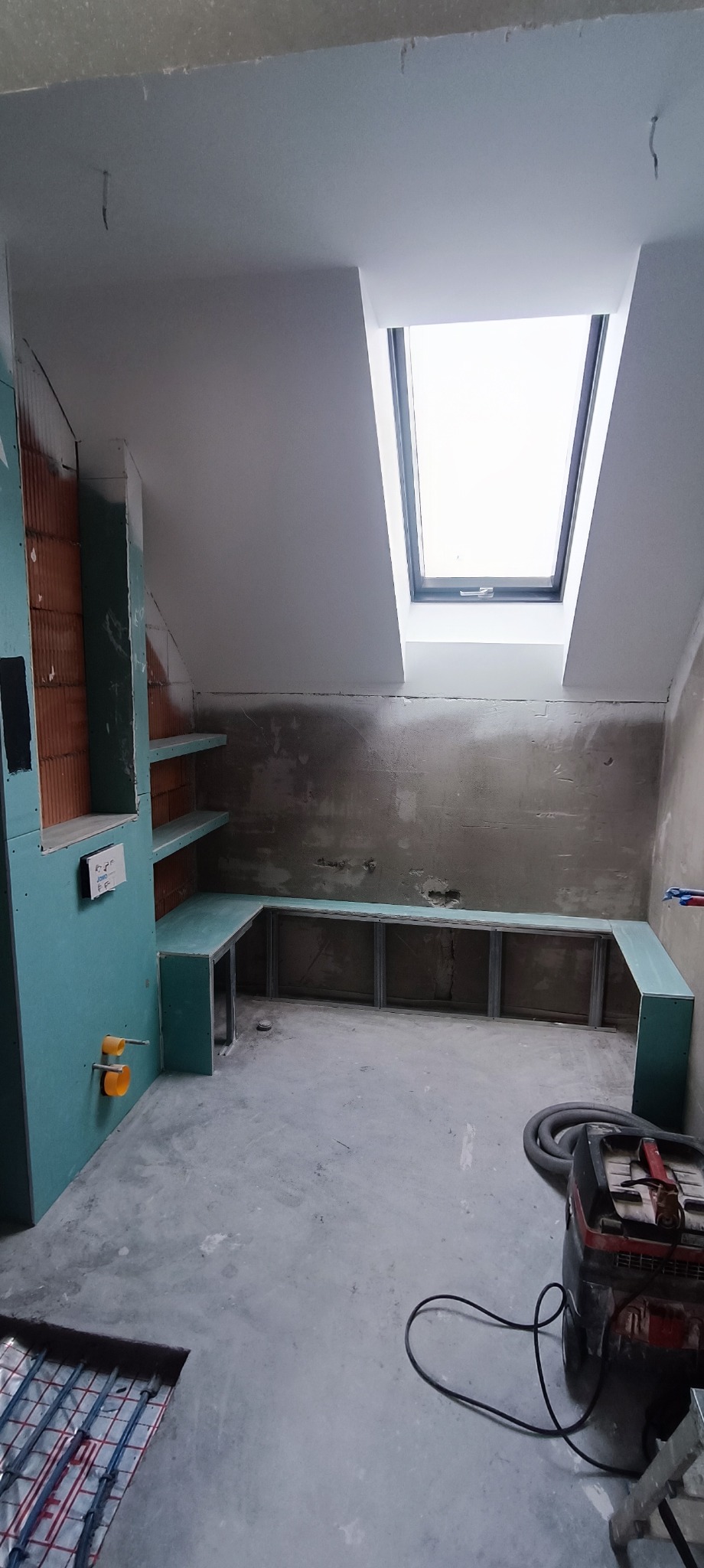 Poddasze w trakcie prac wykończeniowych: zabudowa z płyt kartonowo-gipsowych pod skosem, widoczne profile i fragment instalacji ogrzewania podłogowego, okno dachowe w suficie.