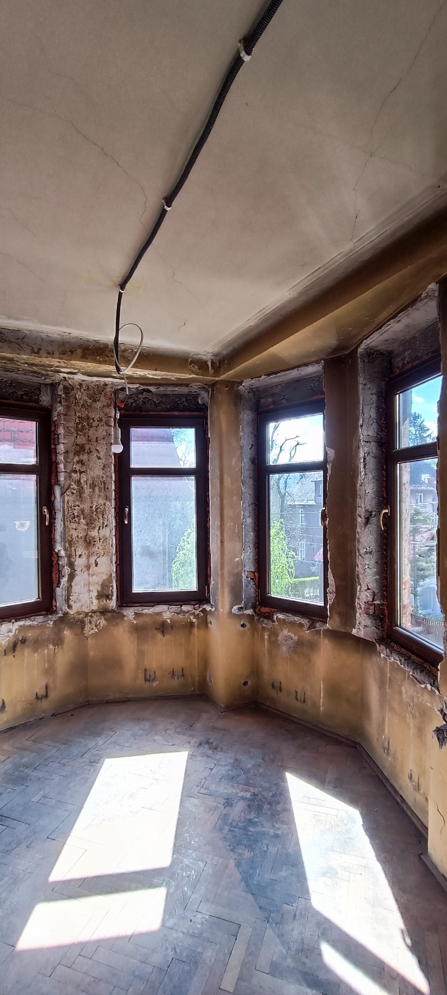 Pokój w trakcie remontu: odsłonięta cegła wokół okien, częściowo usunięta stara farba, widoczne przewody elektryczne na suficie, parkiet z wyraźnymi śladami użytkowania, wpadające promienie słoneczne.