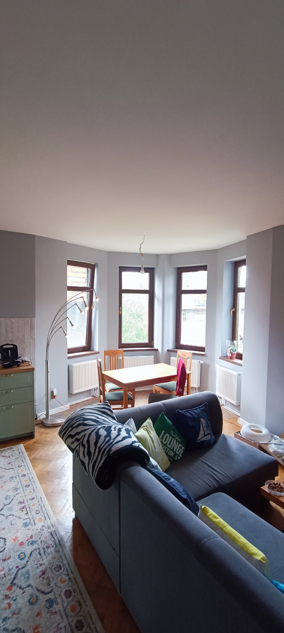 Salon po odświeżeniu: szare ściany, drewniana podłoga, stół z krzesłami przy oknach, szara sofa z poduszkami i wzorzystym pledem.