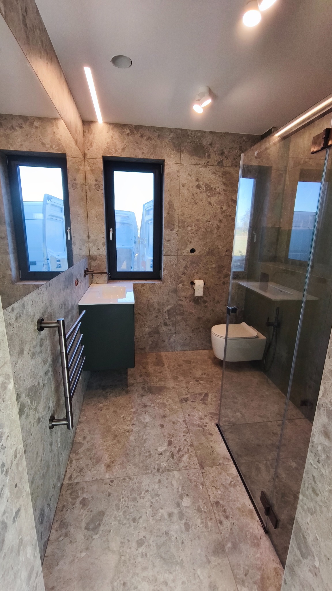 Wykończona łazienka z marmurowymi płytkami, kabiną prysznicową ze szkła, podwieszaną toaletą i zieloną szafką pod umywalką. Widoczne okna z odbiciem samochodów.