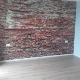 Wnętrze pokoju z fototapetą imitującą starą, czerwoną cegłę na ścianie, widoczne gniazdka elektryczne i jasna, drewnopodobna podłoga.